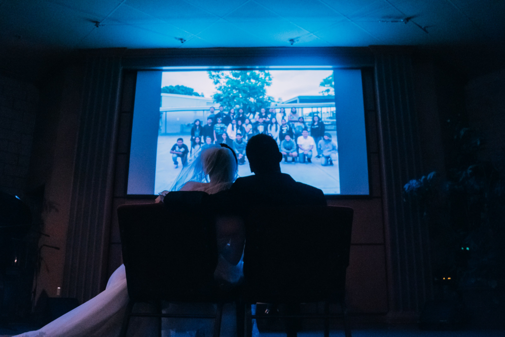 The bride and groom sit quitely to watch their students congratulate them on video